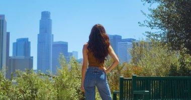 Parkta duran kadın gökdelenlerin tadını çıkarıyor. Kız saati bulanık şehir manzarası