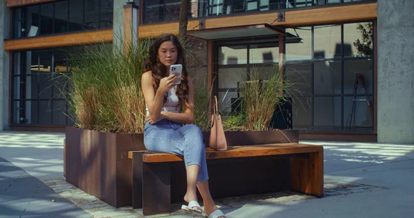 아시아인 브라운 렛 이 시청 벤치에 핸드폰을 놓고 앉아 있습니다. 모바일 기기를 사용하는 소녀. — 스톡 사진