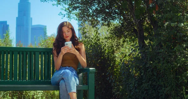 여자들은 벤치에 핸드폰을 놓고 앉아 있는 고층 건물의 모습을 즐긴다. 아시아 소녀는 자연을 진정 시킨다 — 스톡 사진
