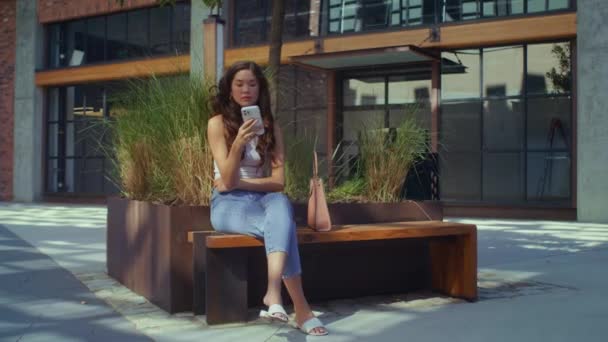 Азиатская брюнетка сидит с телефоном на городской скамейке. Девушка с помощью мобильного гаджета. — стоковое видео