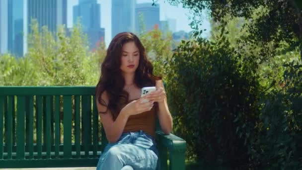 Asiatisk tjej som sitter på parkbänken och håller i telefonen. Kvinna ringa telefonnummer utomhus — Stockvideo