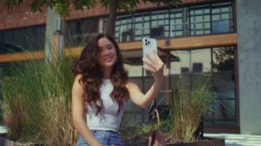 Asyalı kız kenarda otururken kendi fotoğrafını çekiyor. Telefon kamerasında poz veren kız
