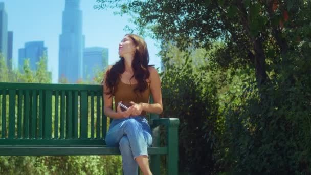 Dame profiter de gratte-ciel vue assis avec téléphone sur le banc. Asiatique fille relax nature — Video