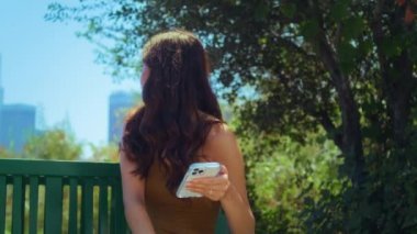 Asyalı kadın bulanık şehir manzarasına bakıyor. Parkta akıllı telefon tutuyor.