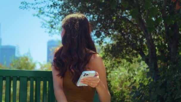 Asiatisk kvinna ser tillbaka på suddig stadsbild håller smartphone i parken närbild — Stockvideo
