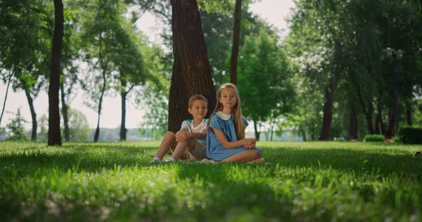 Los hermanos sonrientes juegan en la hierba bajo el árbol. Niños felices sentados en el césped. —  Fotos de Stock