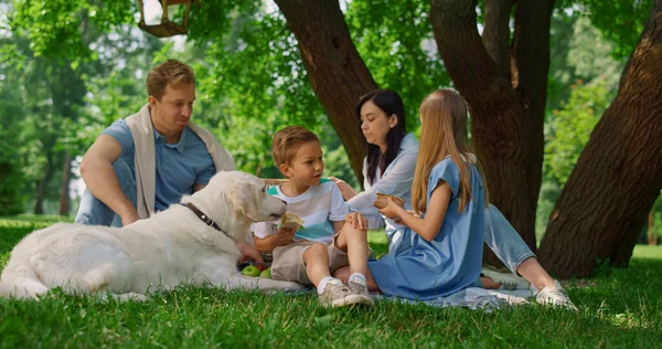 Lycklig familj mata vit hund på picknick. Människor lever roligt på grönt gräs. — Stockfoto