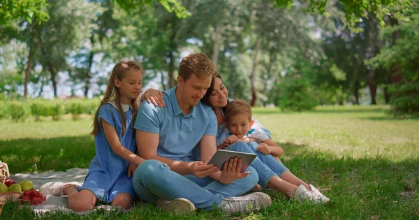 Молода сім'я сидить з таблетками на сонячному парку. Щасливі люди використовують ноутбук на відкритому повітрі — стокове фото