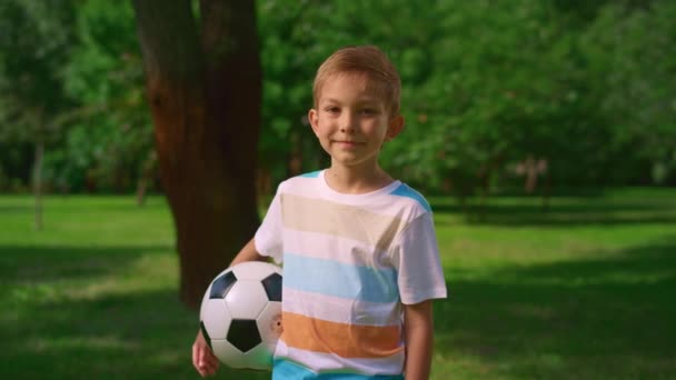 Een klein kind met een bal die glimlacht naar de natuur. Portret van een leuke sportman in het park. — Stockvideo