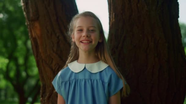 Fröhliches kleines Mädchen posiert am Baumstamm. Lächelndes Kind mit Flechtblick in die Kamera. — Stockvideo