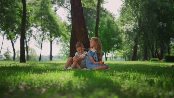 Crianças felizes sentam-se perto da árvore na grama verde. Menina bonito menino jogando na natureza. — Vídeo de Stock
