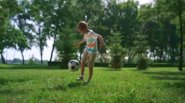 Sportif çocuk parkta futbol egzersizi yapıyor. Genç sporcu güneşli çimlerde topa vuruyor.