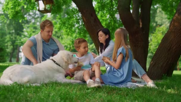 O familie fericită hrănește un câine alb la picnic. Oamenii se distrează pe iarba verde. — Videoclip de stoc