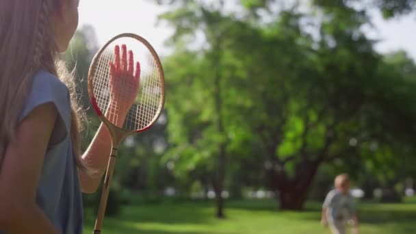 Sıkılmış kız, kardeşi parkta rakete dokunurken badminton oynuyor. Arka görünüm — Stok video