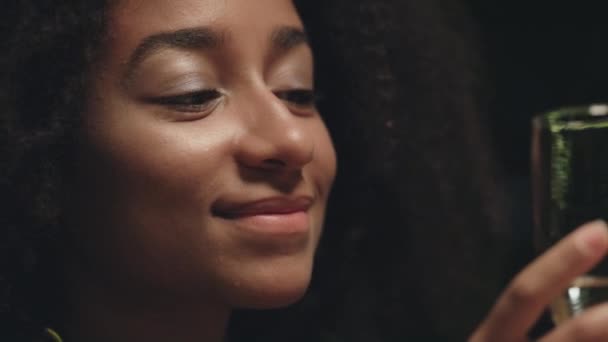 Chica afroamericana bebe vidrio de alcohol. Mujer sexy mira la cámara en luz suave. — Vídeos de Stock