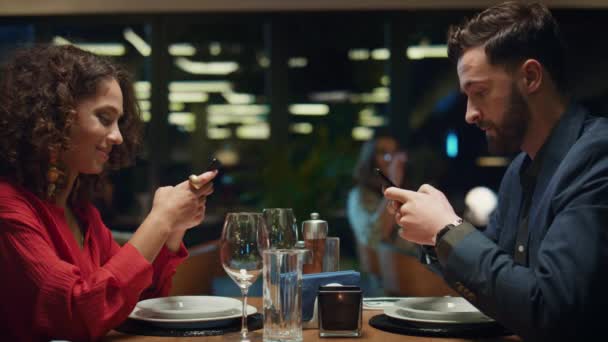 Wieloetniczna para pisząca SMS-y do telefonu komórkowego w eleganckiej restauracji. Koncepcja daty kolacji — Wideo stockowe