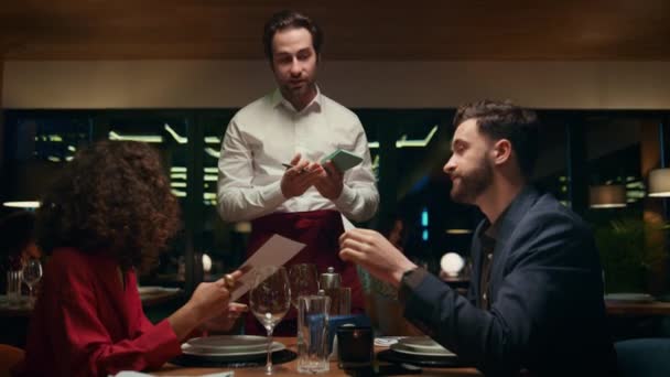 Hübscher Kellner hilft Paar im Restaurant. Multiethnische Liebhaber suchen Menü. — Stockvideo