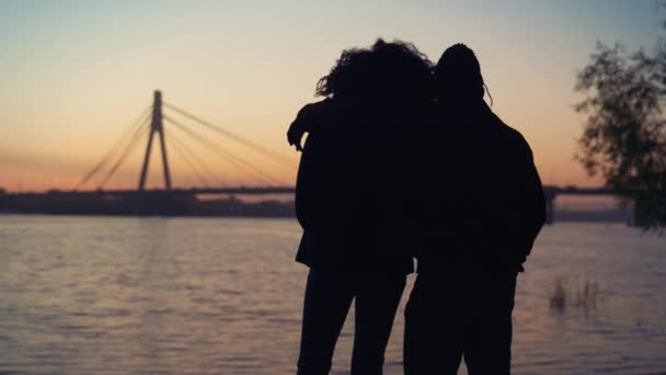 Casal de silhuetas a ver o pôr do sol na vista da ponte do rio. Data romântica ao ar livre. — Vídeo de Stock