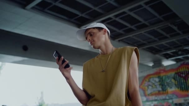 Jovem patinador verificando as mídias sociais no smartphone sentado no parque de skate. — Vídeo de Stock