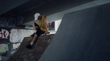 Tarz sahibi bir adam internette cep telefonuyla kaykay parkında grafitiyle internette geziniyor..