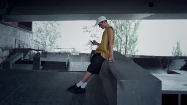 Casual adolescente relajante con teléfono inteligente sentado en skate board en skatepark. — Vídeos de Stock