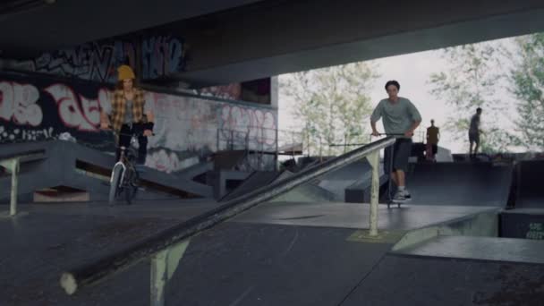 Adolescentes amigos cabalgando juntos en scooter y bicicleta en el parque de skate de la ciudad. — Vídeos de Stock