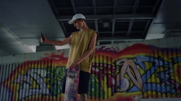Casual skater taking selfie photo on smartphone at skate park graffiti wall. — Stock Video