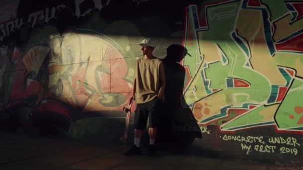 Jeune hipster reposant avec planche à roulettes aux rayons du soleil près du mur de graffité. — Video