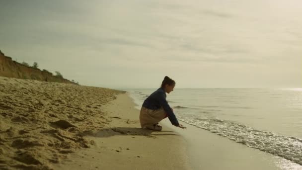 Маленький ребенок касается морских волн на пляже на закате. Милая девушка играет песок в океане. — стоковое видео