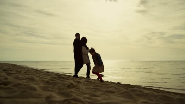 Семья наслаждается закатом пляжа. Игривая девочка бегает вокруг родителей на берегу моря. — стоковое видео