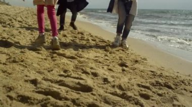Deniz kumu kenarında yürüyen eğlenceli bir aile. Heyecanlı insanların bacakları dışarıda koşturuyor.