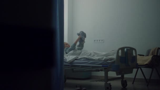 Chora dziewczyna leży sama w szpitalnym łóżku. Zdenerwowane dziecko bawiące się w miękką zabawkę w pokoju kliniki. — Wideo stockowe
