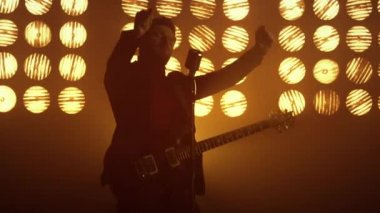 Gitarist sahne ışıkları altında mikrofon şakıyarak eğleniyor. Şarkı söyleyen adam