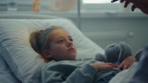 Smutna dziewczyna leżąca w szpitalu przytula zabawkowy portret. Lekarz sprawdza objawy u pacjenta — Wideo stockowe