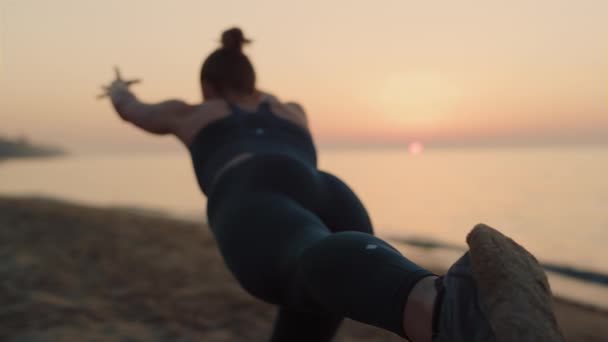 Szczupła dziewczyna ćwicząca wojowniczkę Asanę o zachodzie słońca. Kobieta robi ćwiczenia jogi na piasku — Wideo stockowe