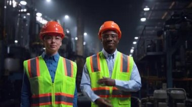 Çok ırklı mühendisler büyük fabrikada güvenlik üniforması giyip gülümsüyor..
