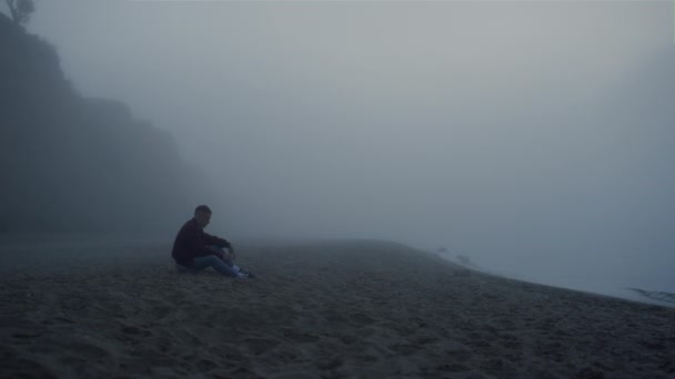En deprimerad kille som sitter på stranden i dimmig morgon. Orolig man röra sand — Stockvideo