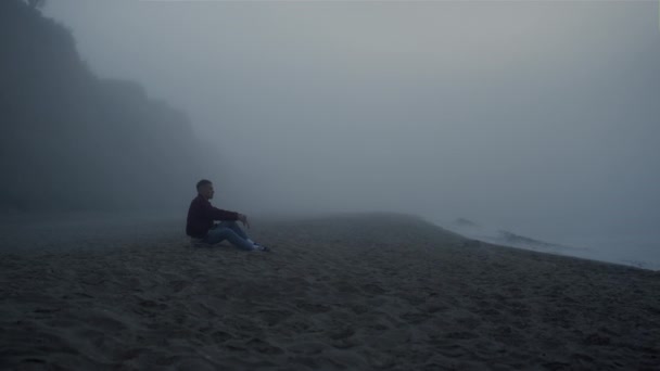 Sad man looking sea landscape in fog. Serious guy sitting on sandy beach — Vídeos de Stock