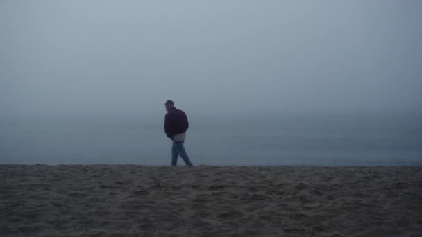 Um tipo sonhador a olhar para o mar de manhã enevoado. Homem perturbado em pé na praia arenosa — Vídeo de Stock