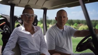 Golf oyuncuları el arabasına bakıyor. Kulüp sahasında oturan çift..