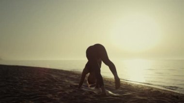 Spor kadını gün doğumunda sahilde yoga yapıyor. Kız esniyor.