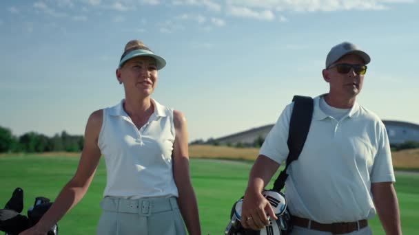 ゴルフ選手のカップルは緑豊かなコースで一緒に歩く。夏におしゃべりする金持ち — ストック動画