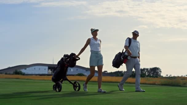 Two golfers walk fairway at sunset. Rich couple carry sport equipment clubs. — Video Stock