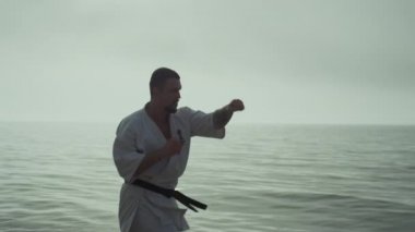 Güçlü bir adam okyanus kenarında tekvando yapıyor. Aktif el hareketleri yapıyor. Spor kaslı adam dışarıda kimono giyerek dinamik yumruklar eğitiyor. Kafkas sporcusu gün batımında sahilde egzersiz yapıyor..