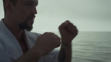 Gün batımı kumsalında el yumrukları antrenmanı için odaklan. Aktif bir adam akşam güneşinin yumuşak ışıklarıyla dışarıda egzersiz yapıyor. Karate sporcusu doğada dövüş sanatları uyguluyor. Fiziksel güç kavramı.