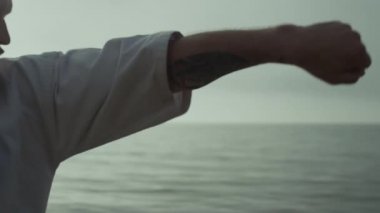 Focused athlete practicing powerful karate punches at sunset outdoor close up. Bearded middle age sportsman making fighting attack wearing kimono near sea waves. Strong man training martial arts.