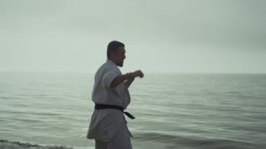 Karate dövüşçüsü gün batımında deniz kıyısında dinamik yumruklar yapıyor. Atletik adam sahilde dövüş teknikleri uyguluyor. Sakallı sporcu antrenman atağı yumuşak akşam güneşinde açık havada. 