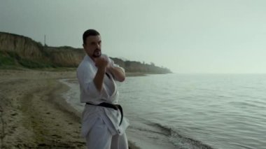 Güçlü sakallı dövüşçü, kumlu sahilde yumruk yumruğa dövüşüyor. Yakından. Sakin denizin yanında karate yapmaya odaklanmış bir sporcu. Tekvando sporcusu dışarıda dövüş tekniği eğitimi alıyor. Spor konsepti.