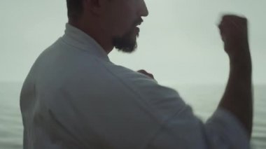 Active sportsman training hands fighting exercises on seacoast close up. Energetic athletic man exercising taekwondo at sunset outdoors. Strong bearded fighter practicing martial technique on nature.