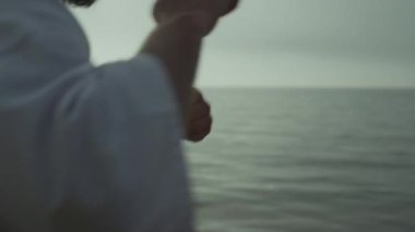 Tattooed man honing fighting skills training punches standing beach at sunrise close up. Active sportsman fists practicing karate attack outdoors. Strong bearded guy exercising in kimono near ocean. 